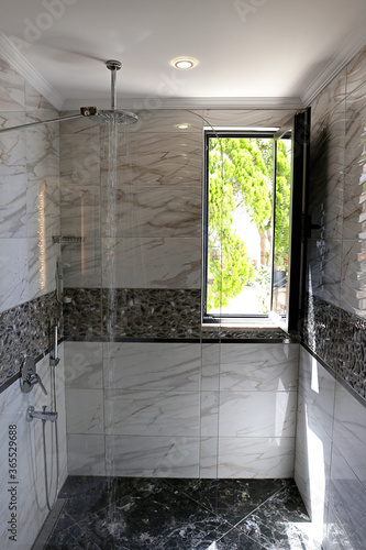 Bathroom filled with natural light through an open window. Shower head mounted to the ceiling with running water. Modern framless fixed glass panel, marble textured tile. Copy space, background. photo