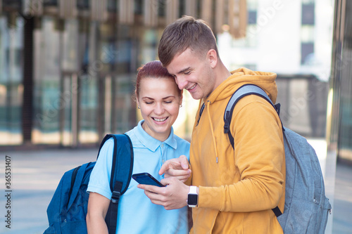 Beautiful young couple, happy students with backpacks standing outdoors together at campus. Guy or boy showing his cell mobile smartphone to girl, pointing with finger at phone, smiling, laughing 