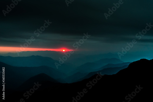 A landscape of the layers of hills with the setting sun in the background and dark cloudy sky above