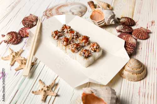 Traditional Japanese Volcano Sushi Roll on carton delivery box with chopsticks. Sea shells and stars on wooden background. Asian dish with raw salmon, flying fish roe (Tobiko) served with Unagi sauce 