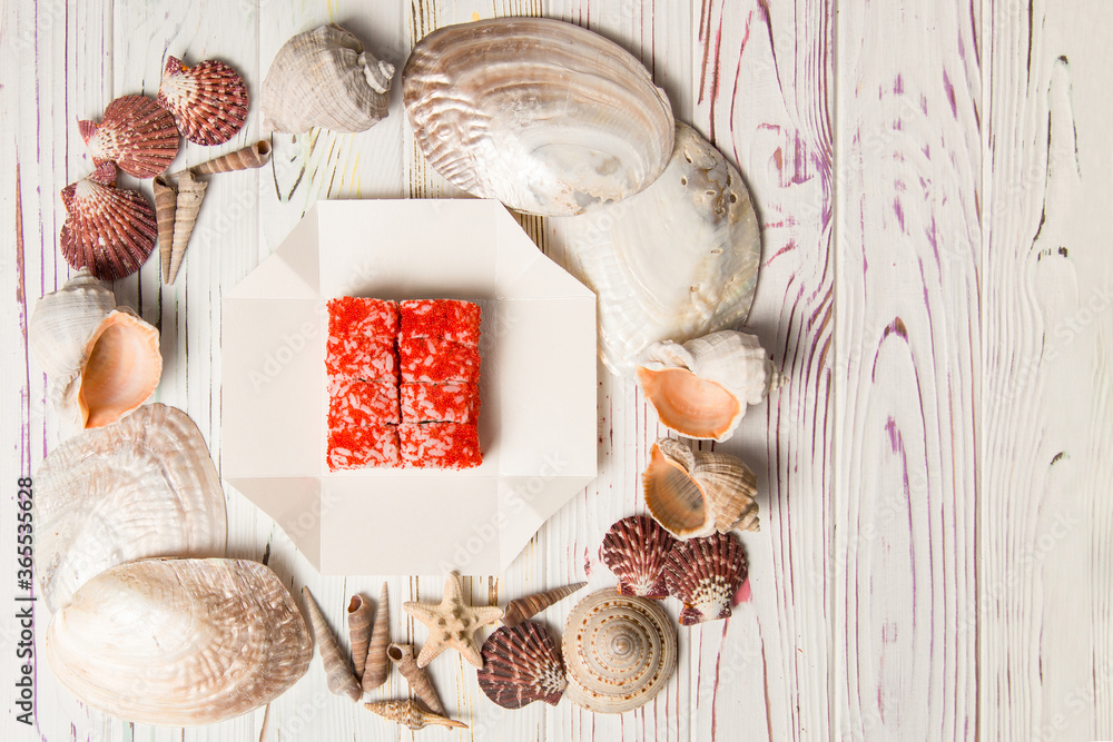 Japanese Sushi in red flying fish roe (Tobiko caviar) isolated on carton delivery takeaway box with Sea shells and stars on white wooden background.
