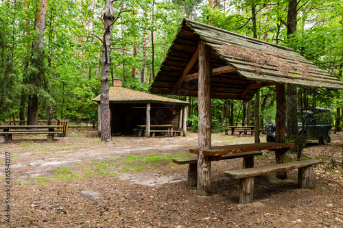 Tourist recreation and rest place in forest. 4x4 offroad car parked next to picnic tables and wooden roof shelters behind trees. Kolacze, Poland, Europe.