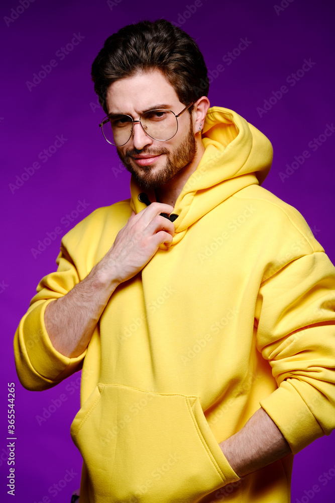 A young man of 25-30 years in glasses and a yellow sweatshirt emotionally poses on a purple background. A young man of 25-30 years in glasses and a yellow sweatshirt emotionally poses on a purple background.