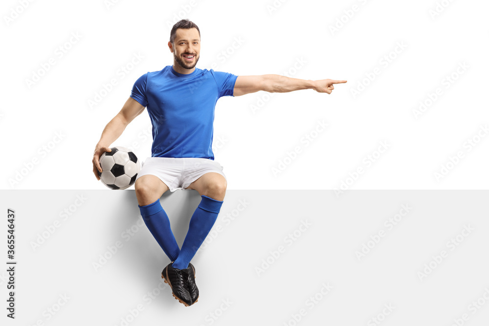 Footballer in a blue jersey with a soccer ball sitting on a blank panel and pointing to the side Footballer in a blue jersey with a soccer ball sitting on a blank panel and pointing to the side