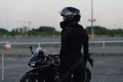 Biker woman in black leather jacket and full face helmet sits near stylish sports motorcycle at urban parking. Traveling and active lifestyle concept. photo