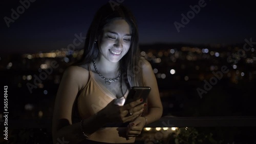 Smiling beautiful young woman wearing sportswear, having break after workout or running, using modern smartphone device at night. Girl typing a sms, texting friends, checking newsfeed in social media
