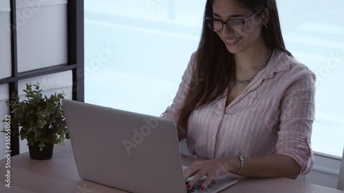 Beautiful young woman manager typing message in online job, surfing web, studying online. Smiling successful business woman in glasses, working remotely on her laptop with big window in the background