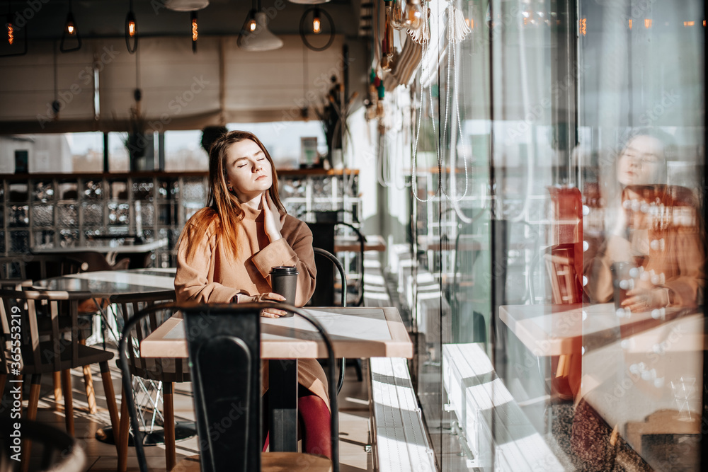 Stylish girl sits in a cafe at the table and drinks coffee. Coffee to go in a cardboard cup. Woman with red hair in a beige warm suit in a cozy atmosphere. Modern interior. Calm and pleasant pastime. Stylish girl sits in a cafe at the table and drinks coffee. Coffee to go in a cardboard cup. Woman with red hair in a beige warm suit in a cozy atmosphere. Modern interior. Calm and pleasant pastime.