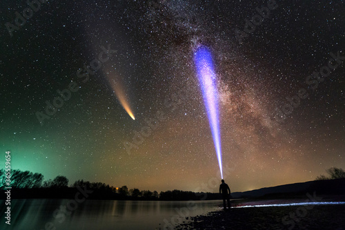 Fototapeta Silhouette of a man with head flashlight standing on river bank lighting a beam of light on Milkyway galaxy and Neowise comet with light tail in dark night sky