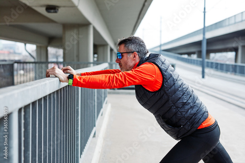 Middle aged caucasian bearded runner stretching legs before running in urban exterior.