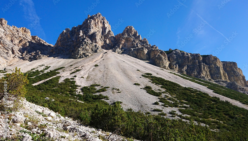 Summer landscape of the Italian Dolomites in the bright rays of the sun. The slope of the white limestone mountain, overgrown with greenery, and the blue sky
