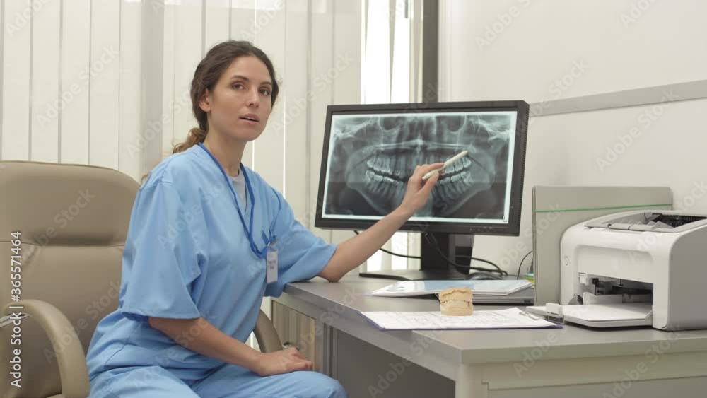 Medium shot of professional female dentist wearing medical overall sitting at desk, holding pen in her hand and examining x-ray image of jaw on computer screen Medium shot of professional female dentist wearing medical overall sitting at desk, holding pen in her hand and examining x-ray image of jaw on computer screen