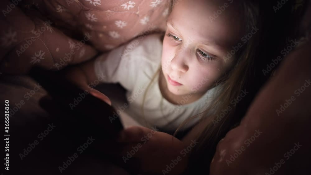 A keen little girl plays on a smartphone at night under a blanket. The child's departure from reality through mobile devices. Child uses secret phone A keen little girl plays on a smartphone at night under a blanket. The child's departure from reality through mobile devices. Child uses secret phone