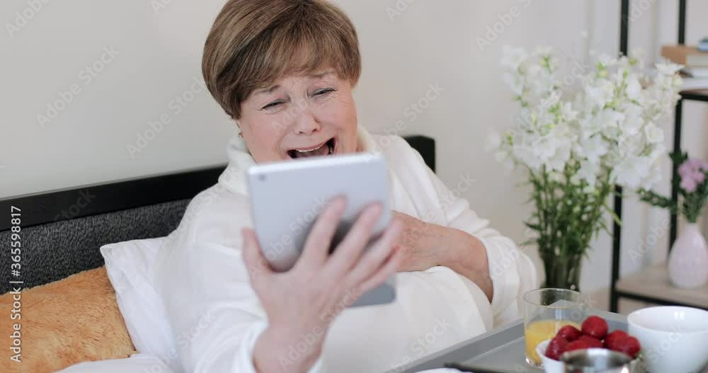 Elderly woman in home rob laughing while looking at screen of her modern gadget. Old female retiree in good mood watching funny video while using tablet and having breakfast in bed. Elderly woman in home rob laughing while looking at screen of her modern gadget. Old female retiree in good mood watching funny video while using tablet and having breakfast in bed.