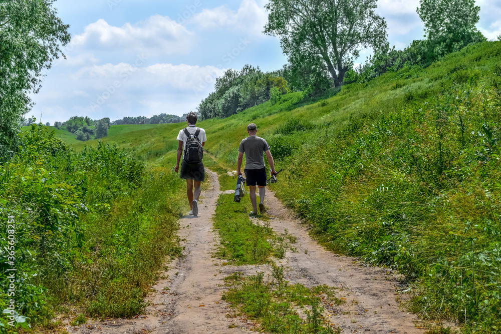 Two fishermen are walking along a meadow dirt road, view from the back. Male hikers with backpack and fishing rods among green nature on a sunny spring-summer day