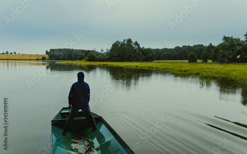 Man standing on fisher boat in pond on moody weather with grass, trees and field. Czech republic