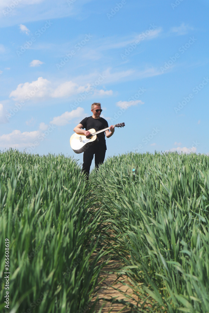 A man in black sunglasses with a guitar in the field. One man sings in nature against a blue sky. Romantic guitarist plays and goes along the path. A man in black sunglasses with a guitar in the field. One man sings in nature against a blue sky. Romantic guitarist plays and goes along the path.