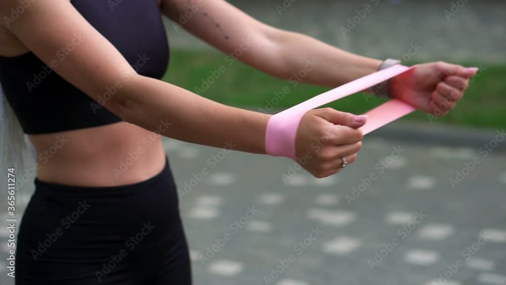 Unrecognizable athletic girl engaged in fitness wearing black sportswear exercising with a rubber band in her hands in a summer park. Girl with small tattoo on hand exercising alone in the morning Unrecognizable athletic girl engaged in fitness wearing black sportswear exercising with a rubber band in her hands in a summer park. Girl with small tattoo on hand exercising alone in the morning