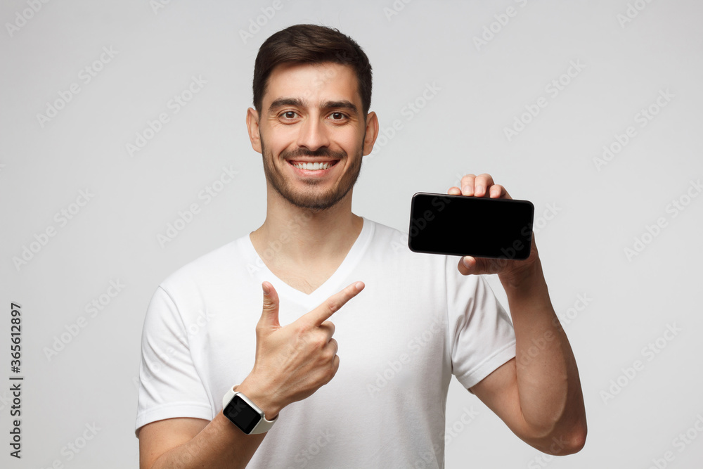 Young handsome guy with short dark hair showing phone with blank screen and pointing to it with finger, isolated on gray background with copy space Young handsome guy with short dark hair showing phone with blank screen and pointing to it with finger, isolated on gray background with copy space