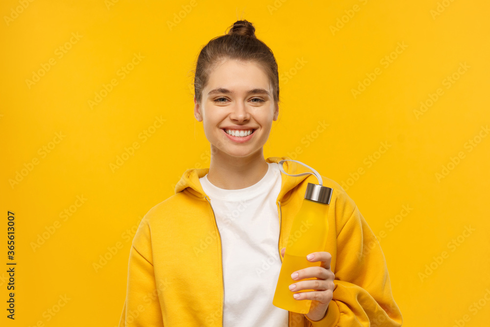 Happy girl in t-shirt and hoodie, holding reusable water bottle in hand, isolated on yellow background Happy girl in t-shirt and hoodie, holding reusable water bottle in hand, isolated on yellow background