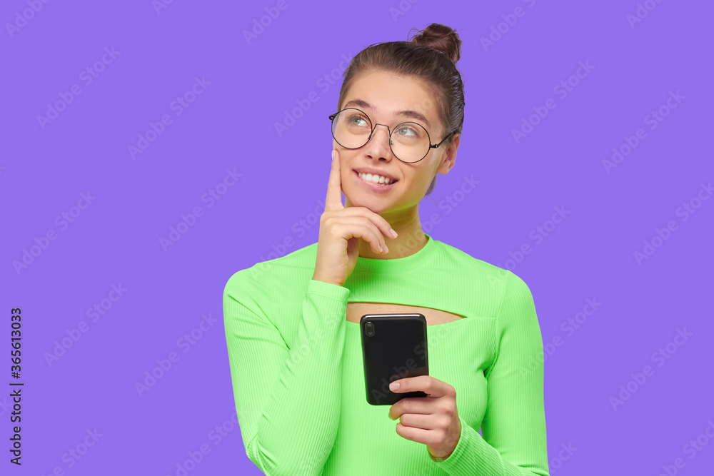 Teen dreraming girl in eyeglasses and headphones around neck, smiling and looking away with phone in hand, isolated on purple background Teen dreraming girl in eyeglasses and headphones around neck, smiling and looking away with phone in hand, isolated on purple background