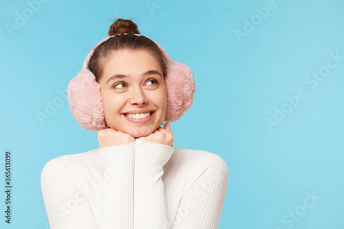 Young girl wearing white turtleneck and pink winter earmuffs looking away with dreamful smile, expecting something good to happen, isolated on blue background