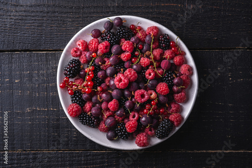 Tasty fresh ripe raspberry, blackberry, gooseberry and red currant berries in plate, healthy food fruit on dark wooden background, top view