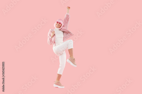 Full length portrait of energetic girl wearing fur coat and colored glasses, jumping up at party, isolated on pink background
