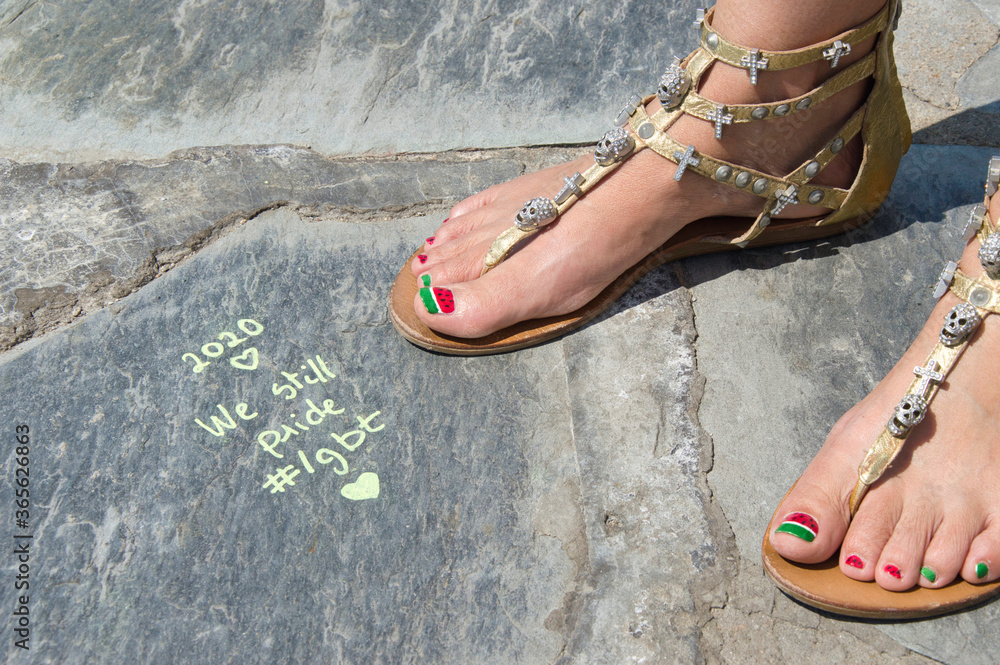 Naklejka premium Cadaqués, Spain, 11 July 2020.A view of a woman's feet in golden sandals reads a sentence about LGTBIQ Pride Day that has been written on a stone floor by the sea.