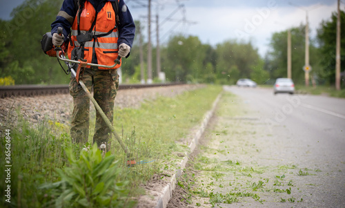 Worker in special protective reflective clothing with a lawn mower in his hands  mows grass with dandelions next to the road and railway tracks. Trimmer in the hands of a man. Cars in the background.
