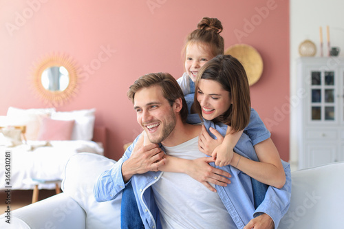 Young Caucasian family with small daughter pose relax on sofa in living room, smiling little girl kid hug embrace parents, show love and gratitude, rest at home together.