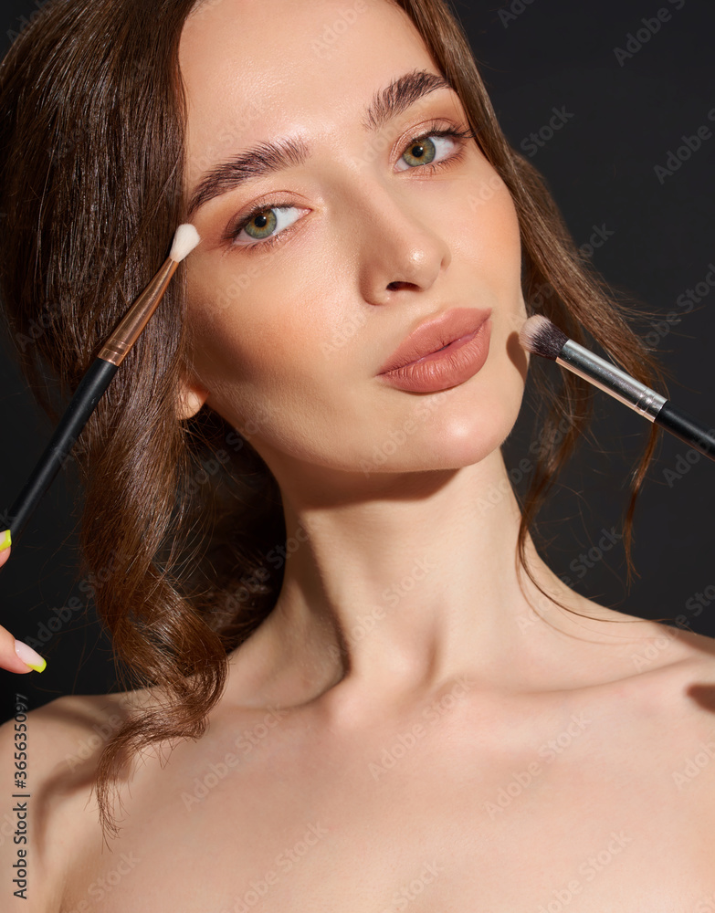 Obraz premium Portrait of the beautiful brunette woman with make-up brushes near face. Girl posing over Gray background. Clean natural makeup.
