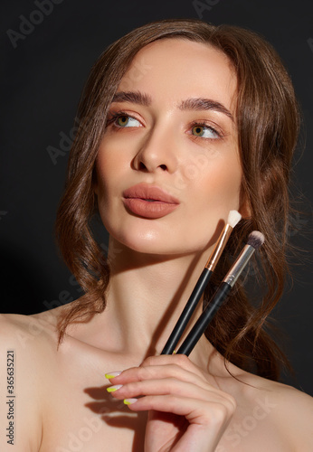 Portrait of the beautiful brunette woman with make-up brushes near face. Girl posing over Gray background. Clean natural makeup.
