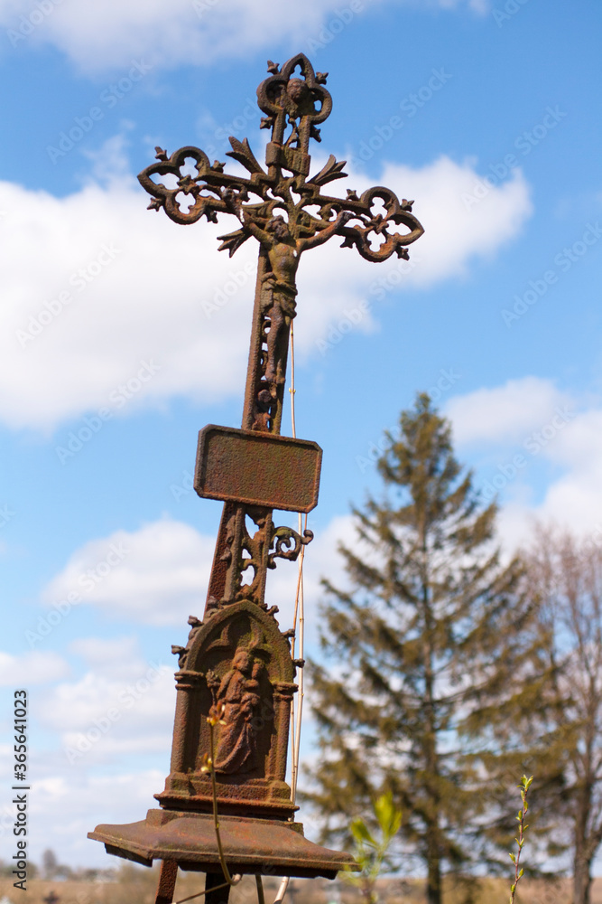 Tilted rusty iron cross in an old cemetery against the sky with clouds.