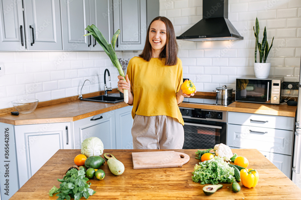 Young and happy woman going to prepare healthy salad with green fresh ingredients in the cozy kitchen. A cheerful girl stands with a leek and pepper in hands and looks at camera