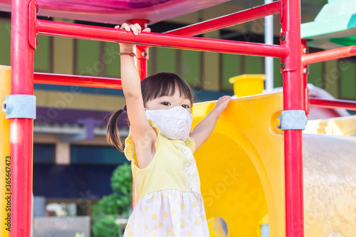 Happy​ Asian​ child​ girl​ wearing​ a​ fabric​ face​ mask​ when​ she​ playing​ a​ toy​ at​ the​ playground.​ Social​ Distance.​ © waridsara