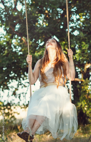 beautiful happy girl with long hair swinging on rope swing on summer nature, young woman enjoy flying in foliage, leisure activity, lifestyle concept