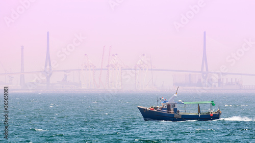 The large suspension bridge of Cadiz in Spain can be seen in the distance in the haze, as well as cranes in the port and ships. In the foreground is a blue fishing trawler.