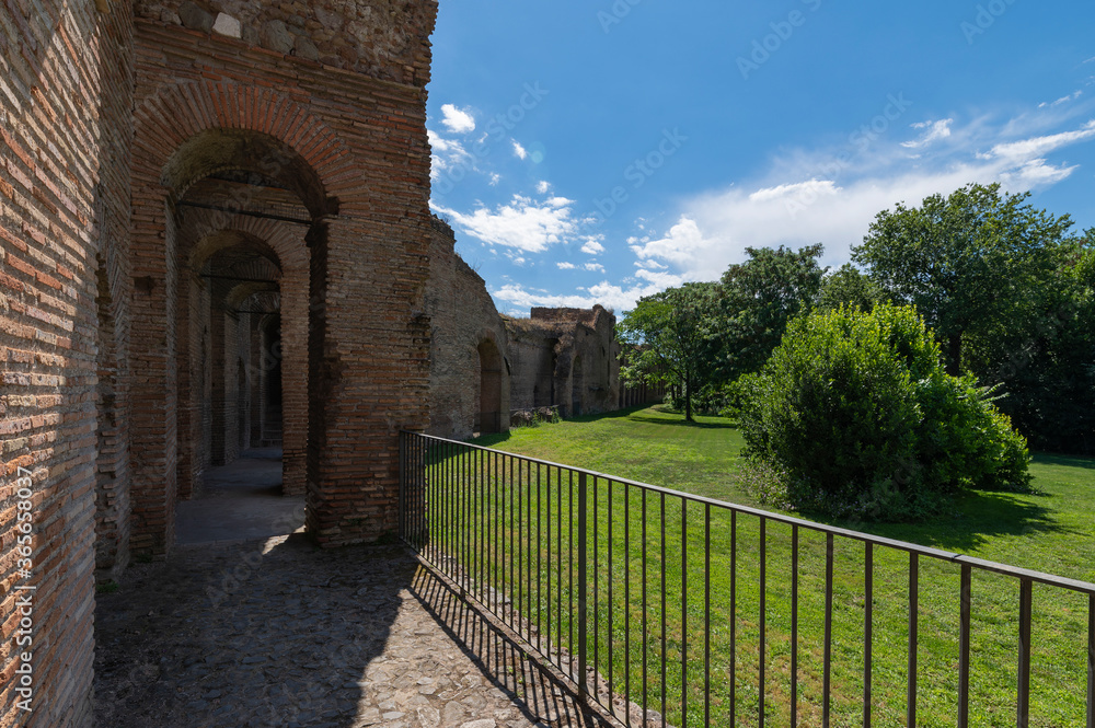The Aurelian Walls of Rome at Porta San Sebastiano seen from inside the city. Brick support arches and cobblestone walkway. The walls border whit a vast lawn with trees. Clear day with clouds. Italy