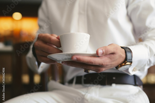 An elegant man in a white suit drinks coffee from a white mug. Hands and mug close -up