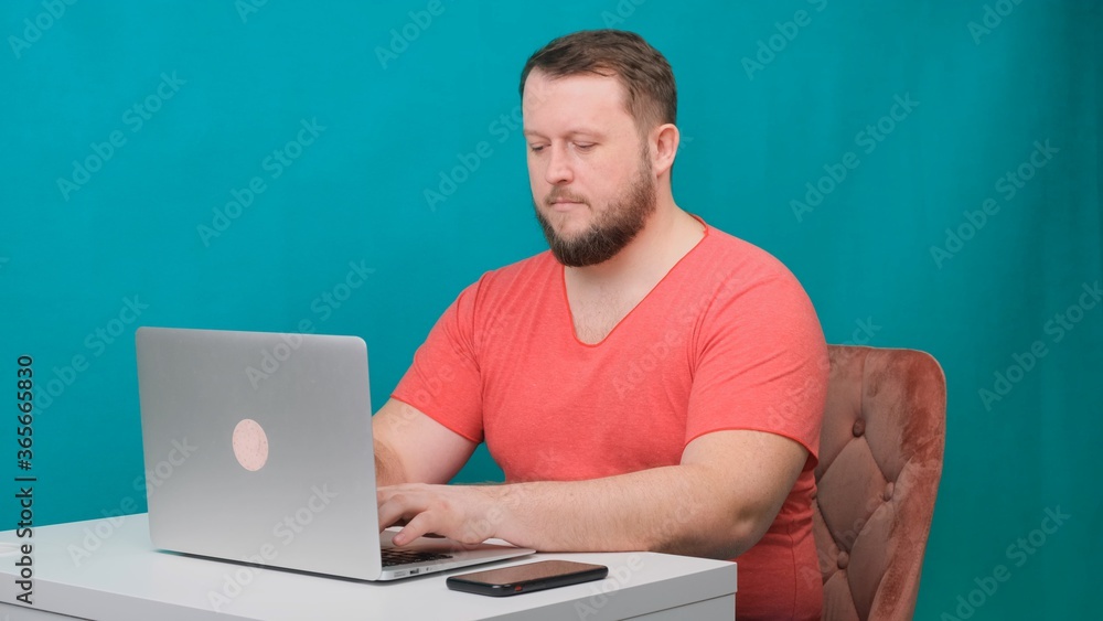 Young happy businessman in a pink t-shirt works on a laptop on a green screen. Portrait of a male talking man looking into his laptop. Man working at his desk in the office. Young happy businessman in a pink t-shirt works on a laptop on a green screen. Portrait of a male talking man looking into his laptop. Man working at his desk in the office.