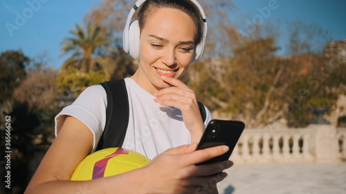 Beautiful sporty girl in headphones with backpack and football happily using smartphone on the way to morning training in city park photo