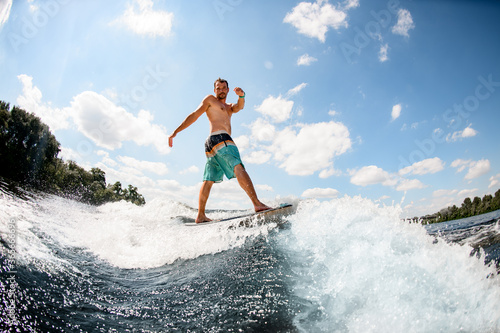 attractive male wakesurfer balancing on board on wave against blue sky