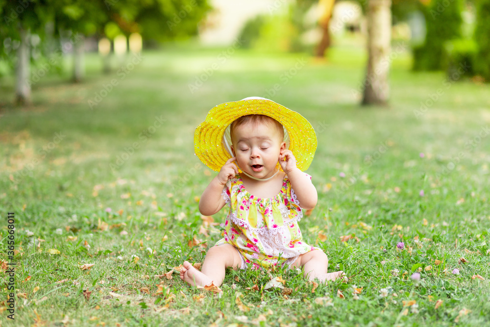 little baby girl 7 months old sitting on the green grass in a yellow dress and hat with her eyes closed and holding her hands to her ears, walking in the fresh air little baby girl 7 months old sitting on the green grass in a yellow dress and hat with her eyes closed and holding her hands to her ears, walking in the fresh air