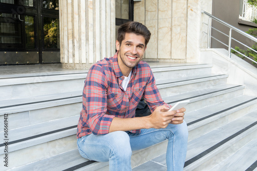 Young handsome guy holding an electronic book with a beautiful smile sitting on the stairs 