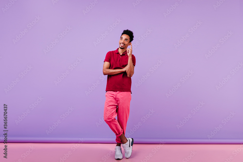 Inspired smiling man in red t-shirt enjoying indoor photoshoot. Handsome guy with short wavy hair standing on purple background. Inspired smiling man in red t-shirt enjoying indoor photoshoot. Handsome guy with short wavy hair standing on purple background.