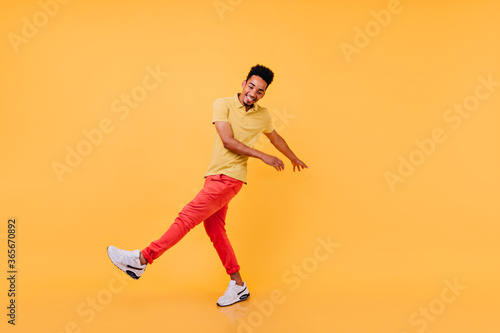 Inspired black guy in bright casual attire funny dancing. Laughing african male model in yellow t-shirt and red pants fooling around in studio.