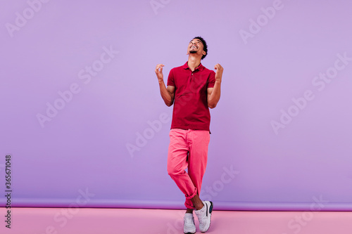 Emotional young man in pink pants standing with legs crossed. Indoor photo of good-humoured guy posing with pleasure on purple background.