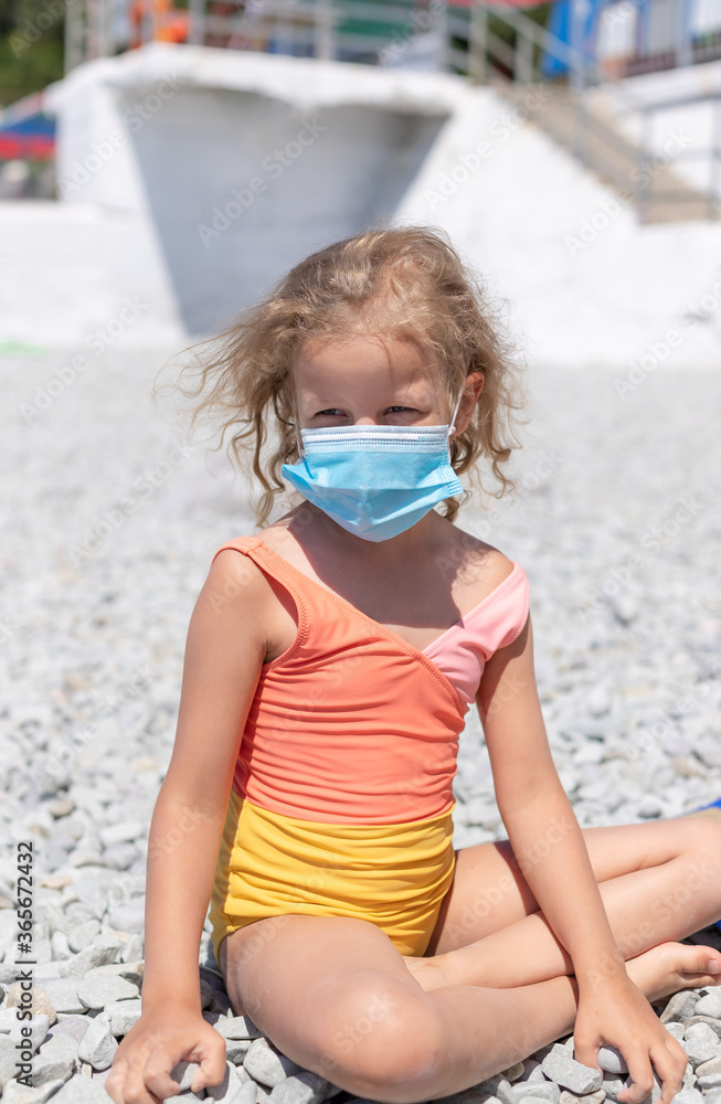 little blond girl in a swimsuit and a protective medical mask on a pebble beach. New rules to wear medical face covering mask at public places due coronavirus COVID 19.vertical image