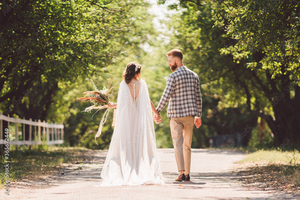 Happy bride and groom holding hands and walking in garden wedding day. Back view of charming stylish newlyweds holding hands while walking in park forest. Path to future life. happy marriage moments Happy bride and groom holding hands and walking in garden wedding day. Back view of charming stylish newlyweds holding hands while walking in park forest. Path to future life. happy marriage moments